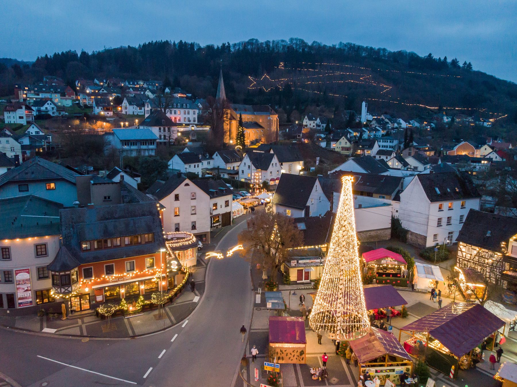 Ein festlicher Weihnachtsmarkt mit einem großen beleuchteten Baum steht im Zentrum einer kleinen Stadt, umgeben von geschmückten Buden und malerischen Häusern. In der Abenddämmerung sind im Hintergrund Hügel mit glitzernden Lichtern und eine Kirche zu sehen.